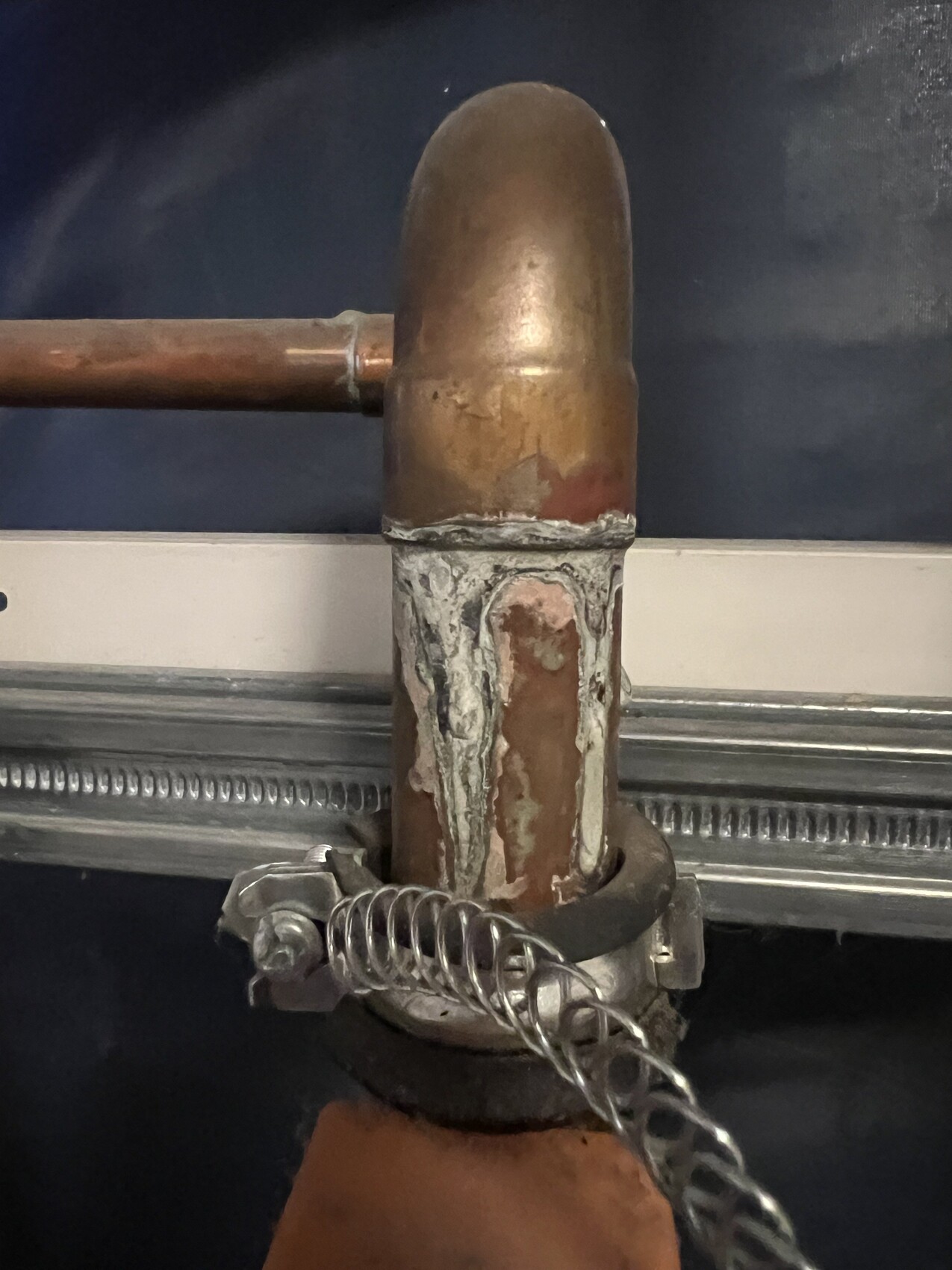 Verrostete Kupferrohrverbindung mit Bogenstück, grauer Dichtpaste und Klemme mit Kette.