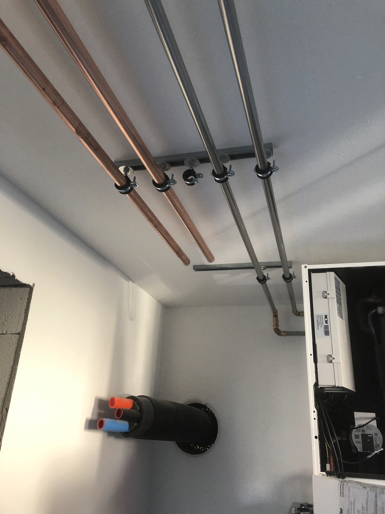 Mehrere Kupfer- und Metallrohre hängen an der Decke, mit Schellen; rechts ein Schaltschrank.