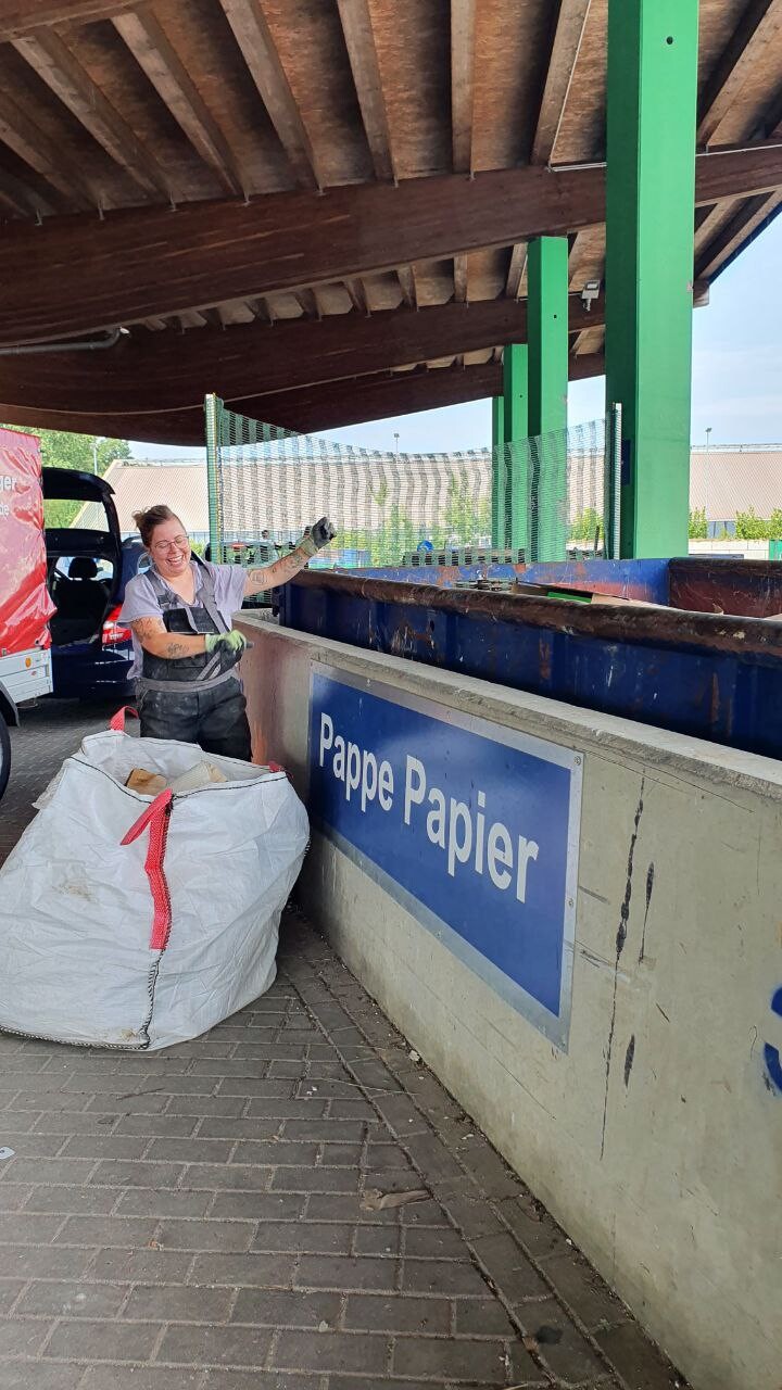 Lächelnde Frau in Arbeitskleidung neben Pappe/Papier-Recyclingbehälter; weißer Sack voller Papier.