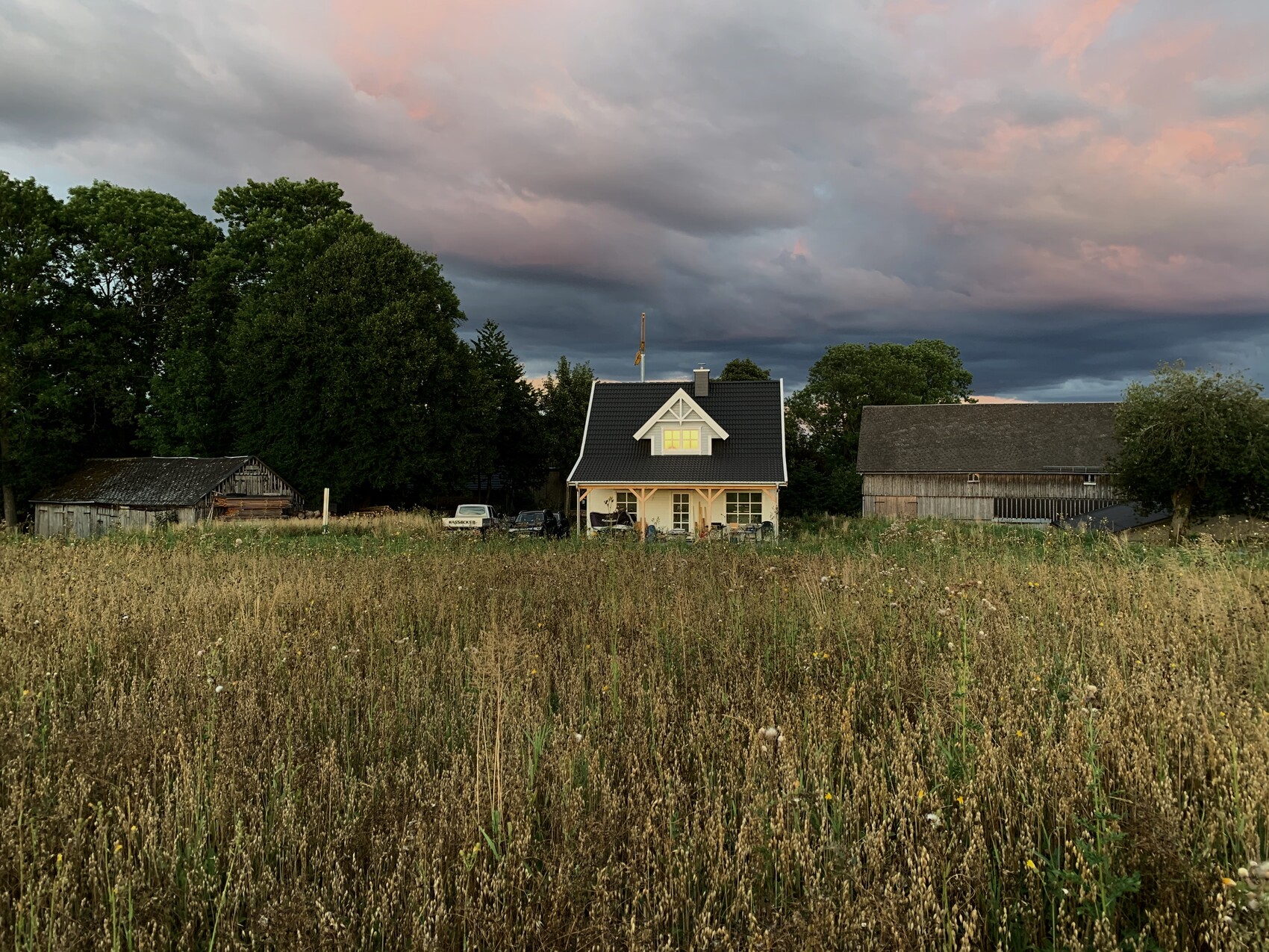 Kleines Haus mit dunklem Dach und Veranda, Scheunen, Bäume, Autos vor dem Haus, Abendhimmel.