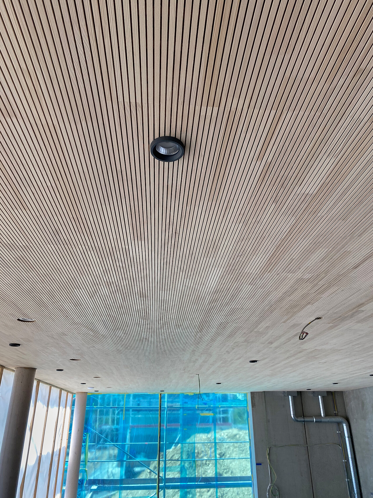 Decke mit parallel verlaufenden Holzlatten; runde Einbauleuchte, Baustelle hinter Glas sichtbar.