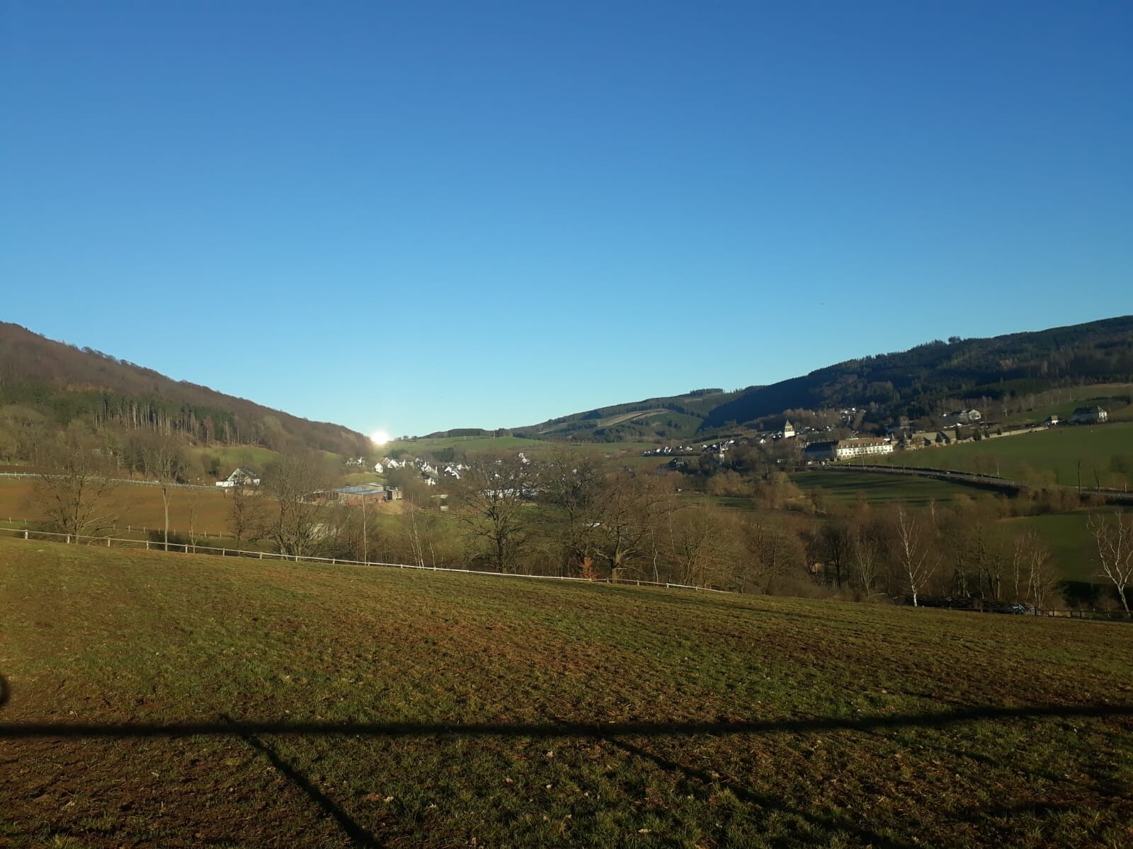 Landschaft mit Feldern, Hügeln und kleinem Dorf im Tal unter blauem Himmel.
