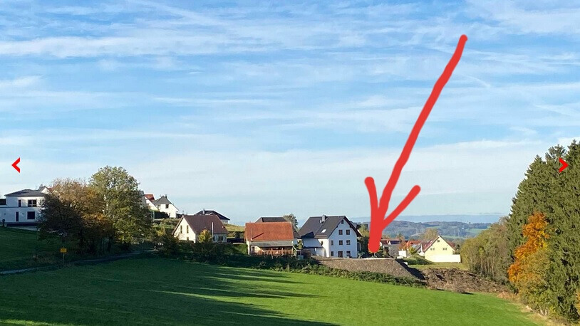 Kleines Dorf mit weißen Häusern am Hang, grüne Wiesen davor; roter Pfeil zeigt auf ein Haus.