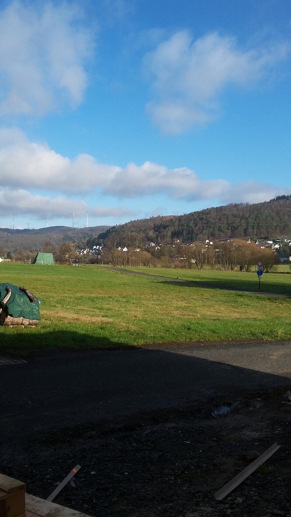 Weite grüne Wiesenlandschaft mit Windrädern am Horizont, Dorf am Hang und blauem Himmel.