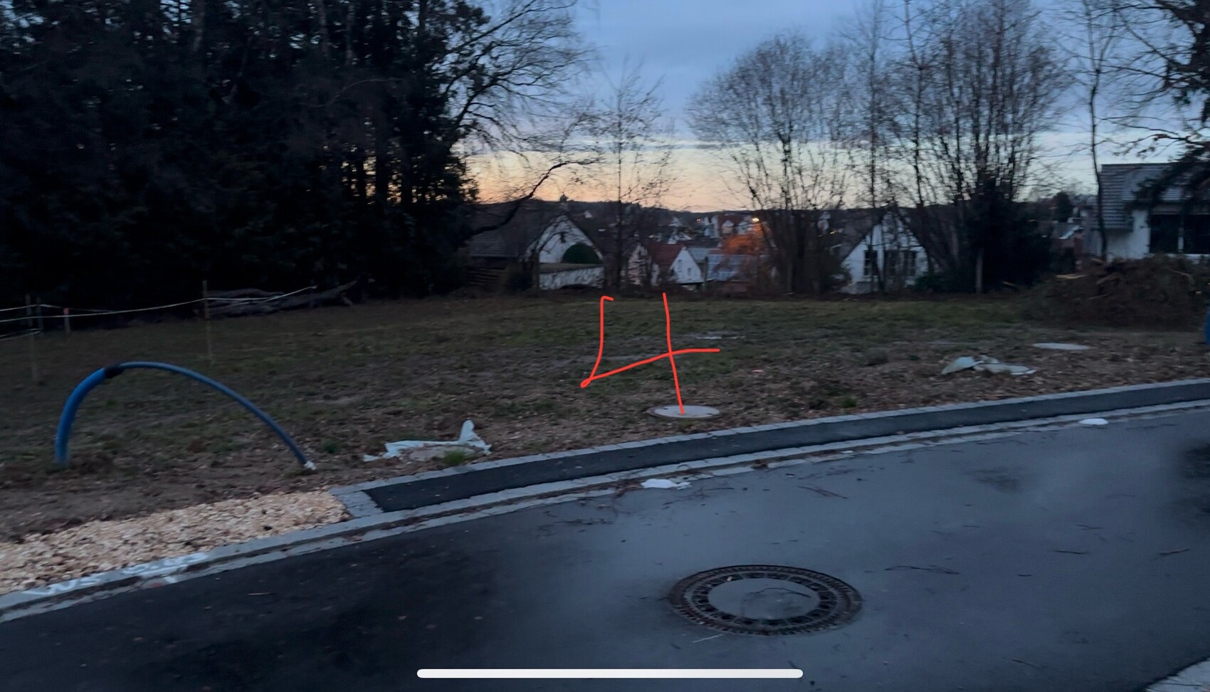Abendliche Straßenseite: nasser Asphalt, blauer Spielbogen, kahle Bäume, Häuser im Hintergrund.