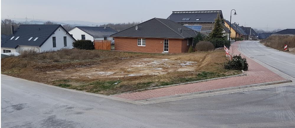 Leerstehendes Baugrundstück an einer Straßenecke mit kleinem braunen Haus im Hintergrund und Gehweg.