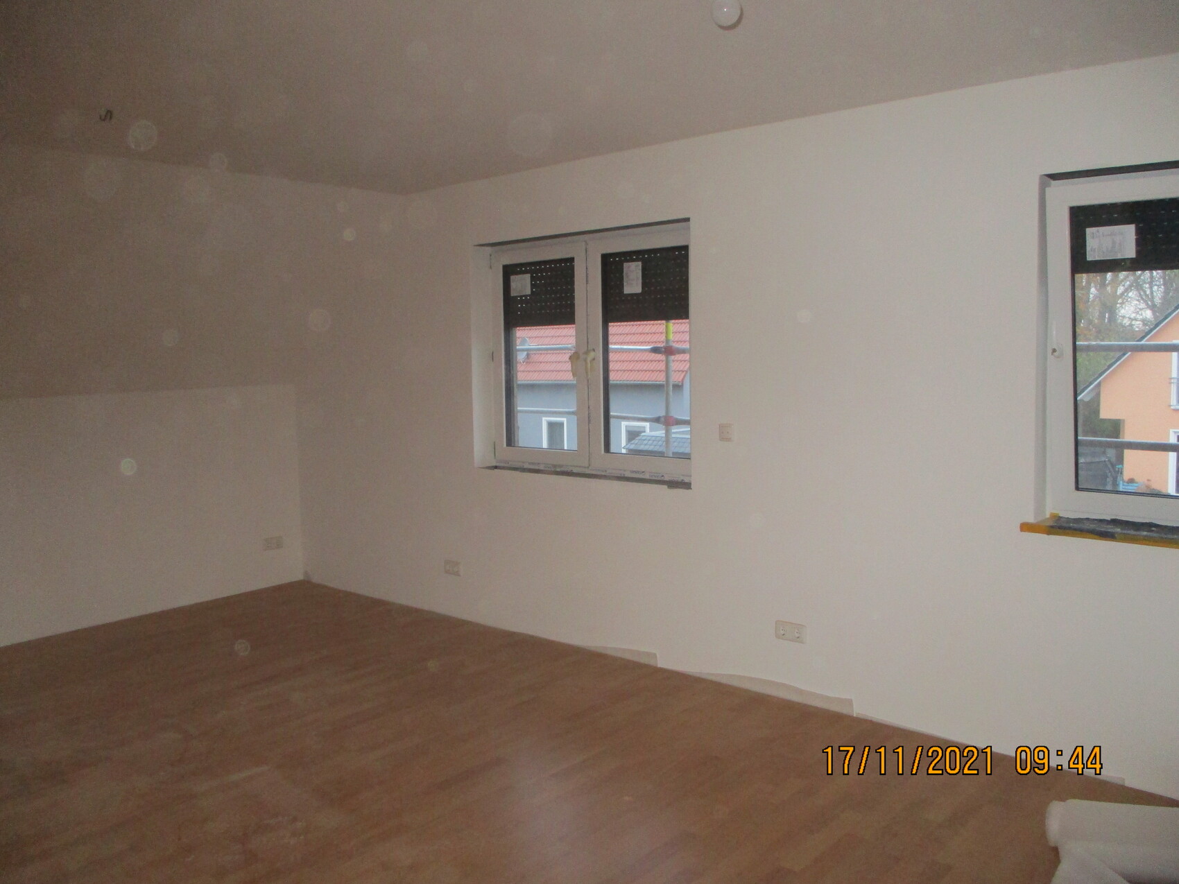 Leeres Renovierungszimmer mit Holzboden, weißen Wänden und zwei Fenstern, Blick nach Nachbarhäusern.