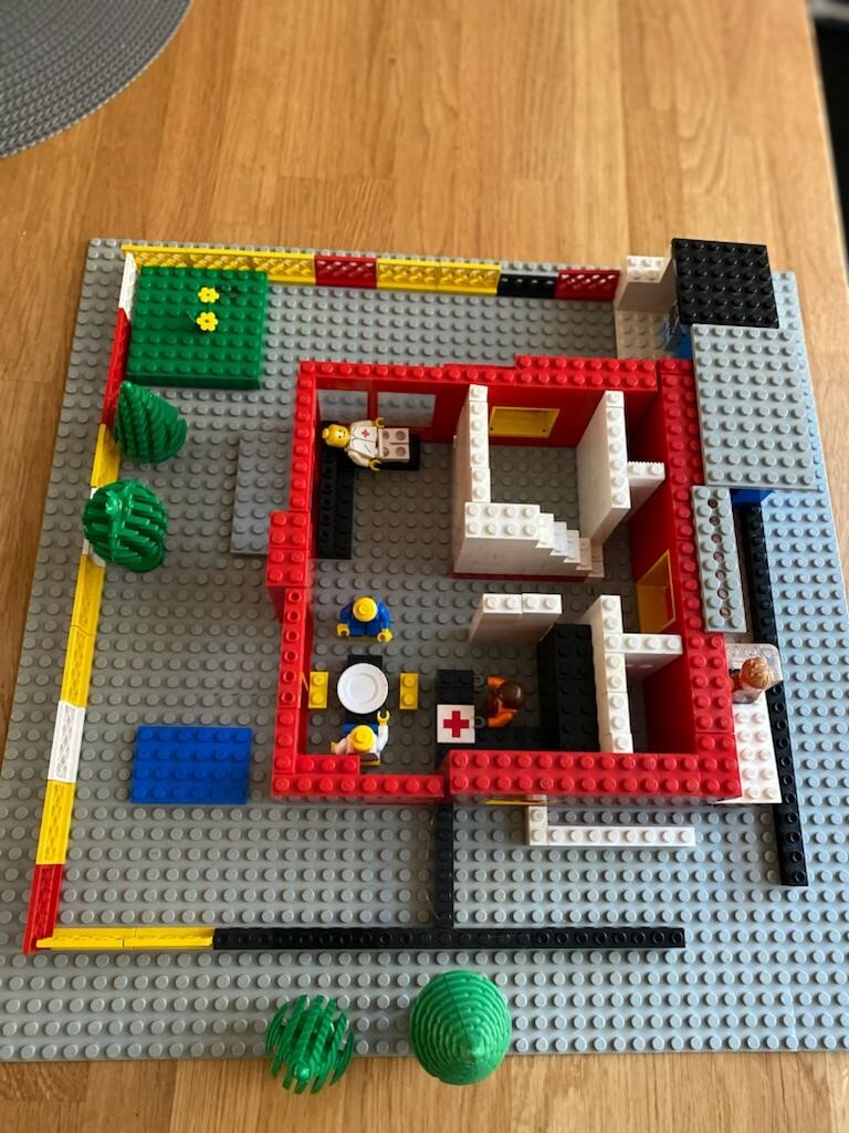 LEGO-Klinikaufbau mit rotem Innenraum, weißen Wänden, Minifiguren und Erste-Hilfe-Kreuz.