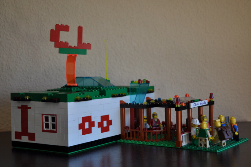 LEGO-Hausmodell mit Minifiguren und Café-Terrasse vor weißem Bau mit rotem Design