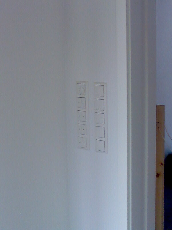 Weiße Wand mit zwei vertikalen Lichtschalterleisten neben Türrahmen, dahinter heller Raum sichtbar.