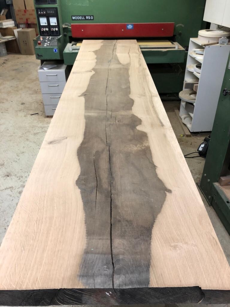 Lange Holzbohle mit dunklem Mittelteil und Riss, in einer Holzwerkstatt neben Maschinen.