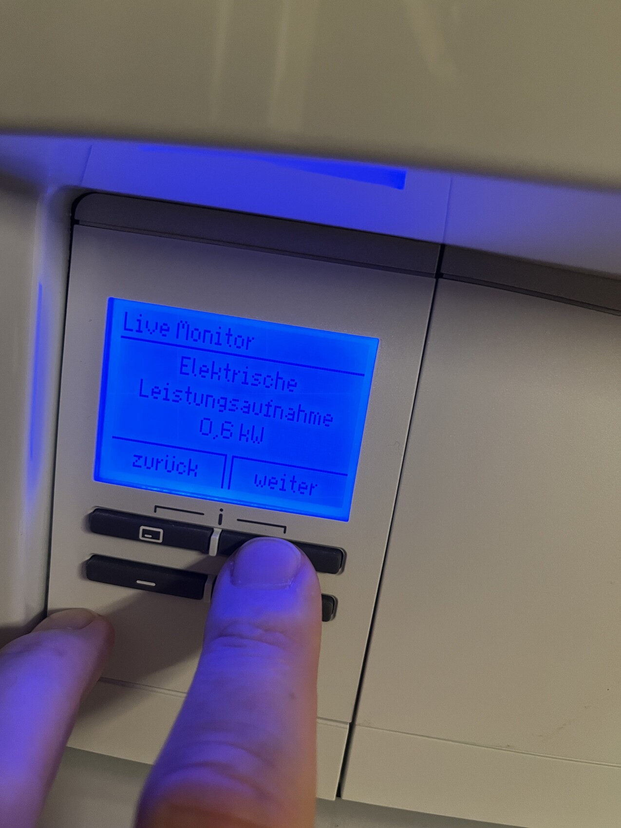 Bedienfeld mit blauem Display: Live Monitor, Elektrische Leistungsaufnahme 0 kW; Finger am Knopf.