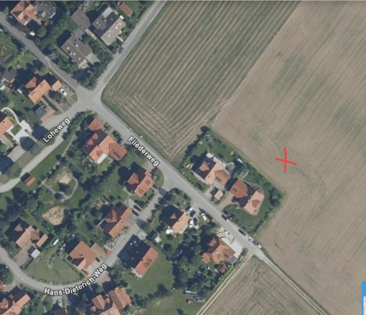 Luftaufnahme eines Wohngebiets mit roten Dächern links, Feld rechts, rotes X markiert.