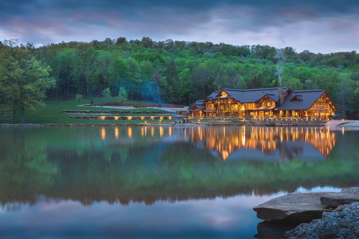 Großes Holzhaus am See mit Lichtern, Wald im Hintergrund und Spiegelung im Wasser.