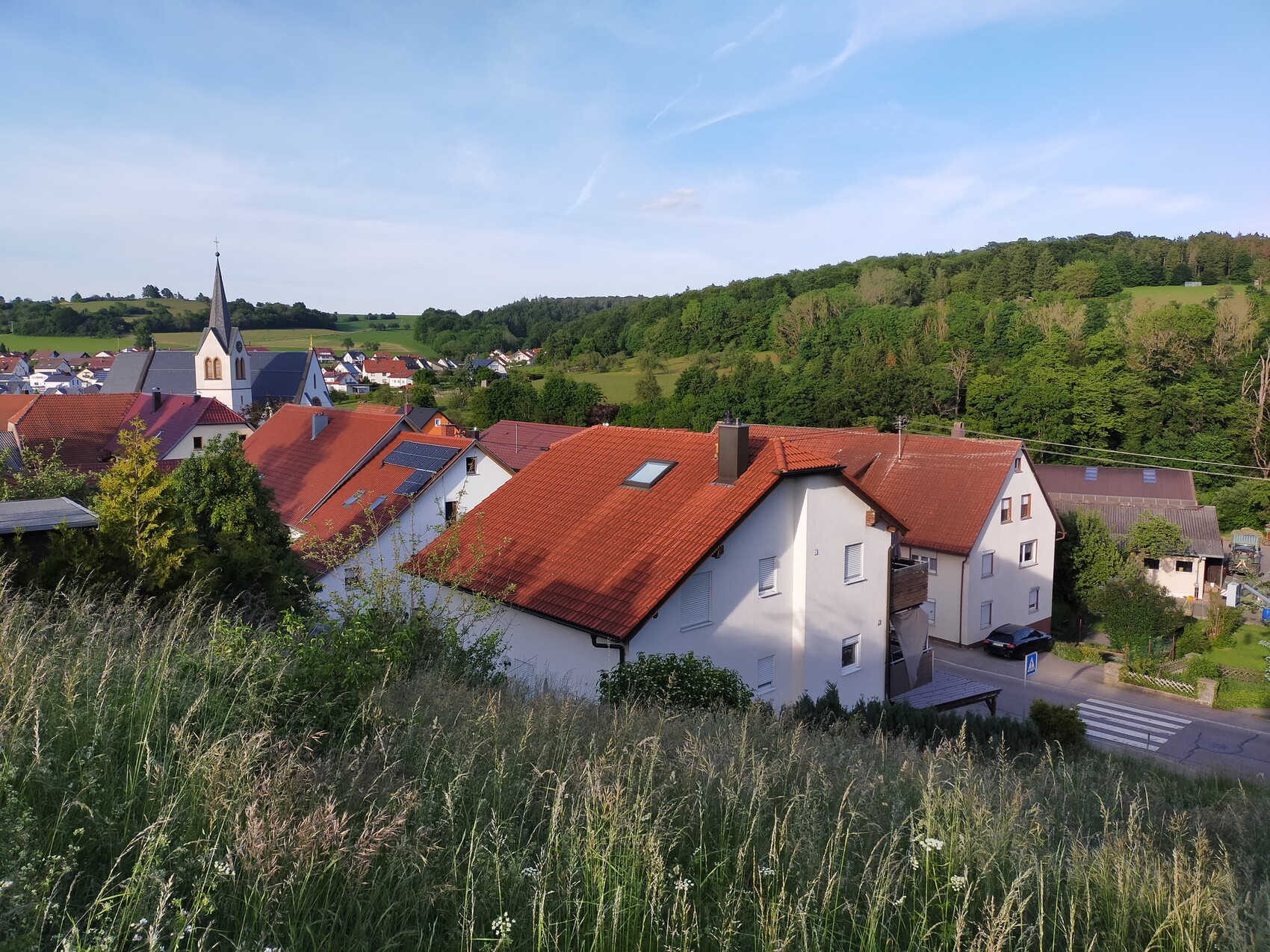Blick auf ein Dorf mit roten Ziegeldächern und Kirchturm, umgeben von grünen Hügeln.