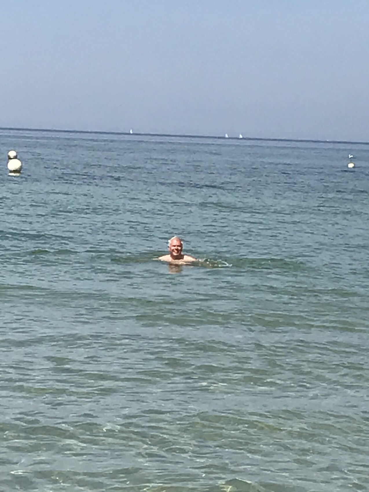Eine Person schwimmt im klaren Meer; Bojen im Wasser und Segelboote am Horizont.