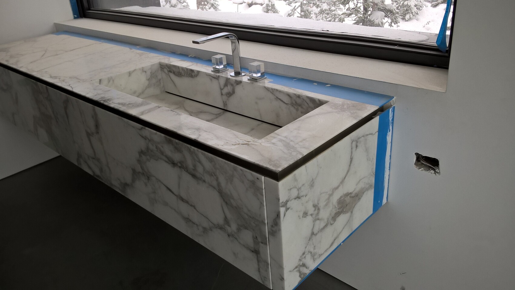 Moderne Marmorspüle mit Chrome-Armatur in Baustellen-Badezimmer; blaue Klebebänder, Schnee draußen.