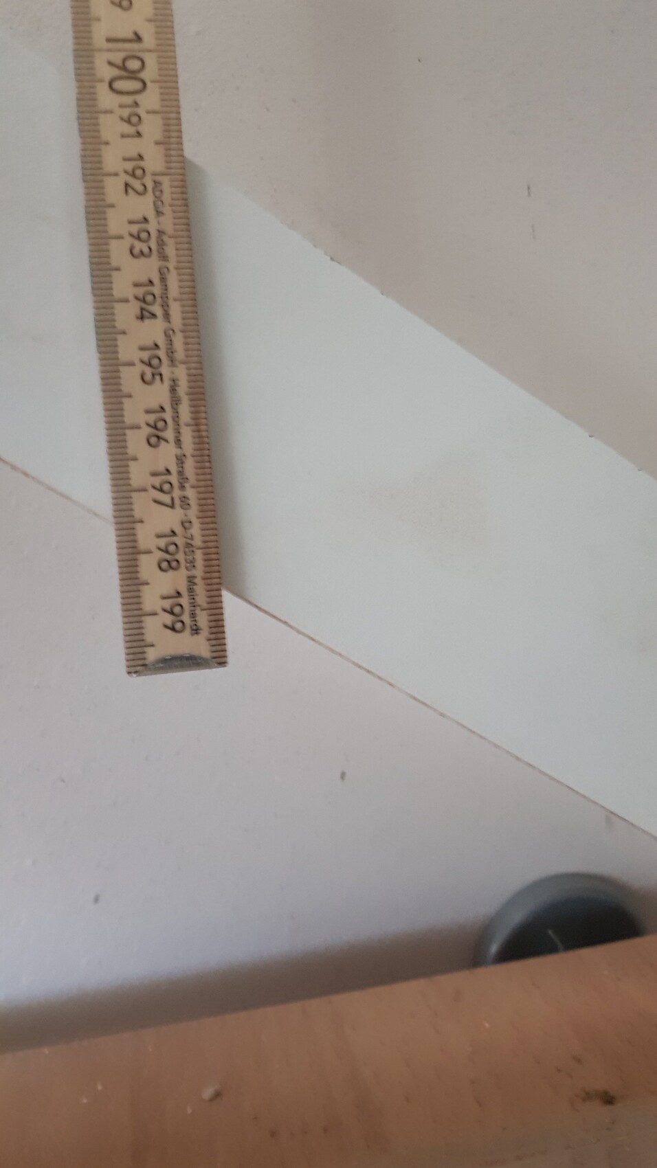 Holzmaßband steht senkrecht an einer weißen Wand; Skala 190–199 cm sichtbar.