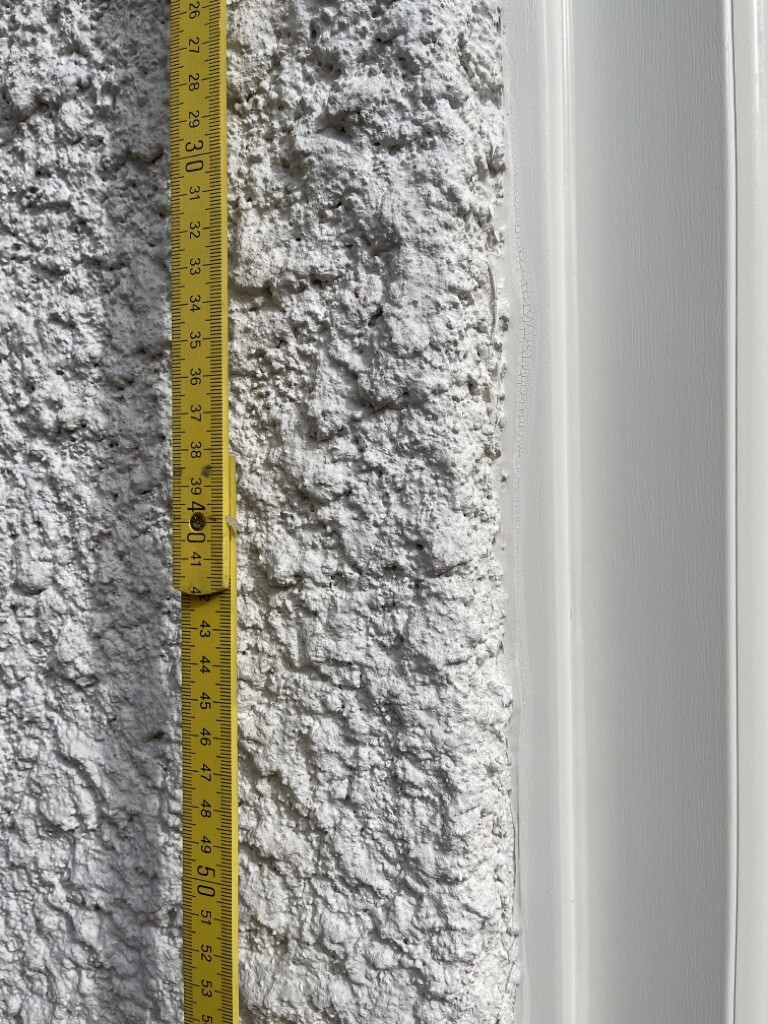 Gelbes Maßband senkrecht an rauer weißer Wand neben glatten Türrahmen.
