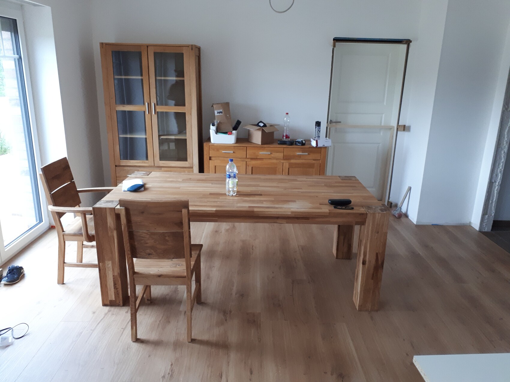 Großer Holztisch mit Stühlen in hellem Raum; Glasvitrine, Kommode und Tür dahinter.