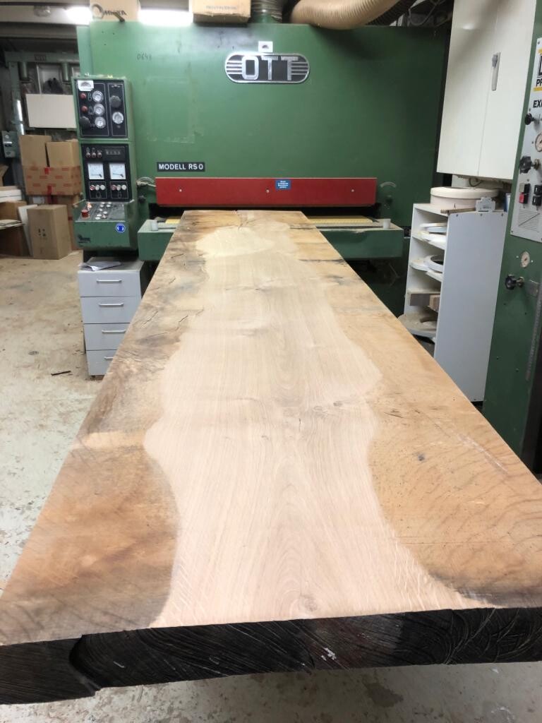 Lange Holzplatte liegt auf einer großen Holzbearbeitungsmaschine in einer Werkstatt.