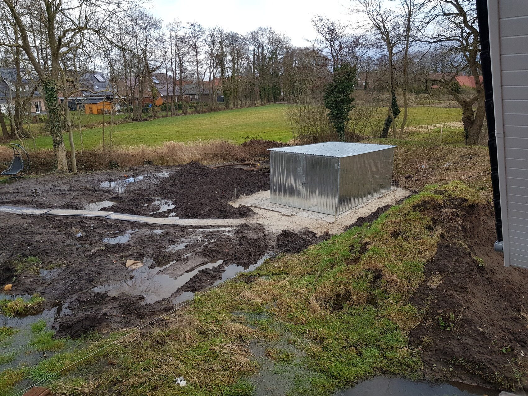 Gartenbaustelle mit silbernem Metall-Schuppen, Schlamm und Pfützen, Hintergrund mit Häusern.
