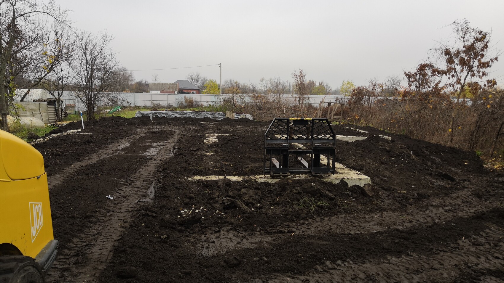 Matschige Baustelle mit gelber Baumaschine links und schwarzem Geruest auf Betonplatte in Mitte.