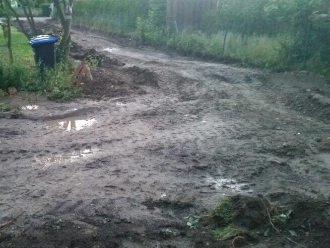 Matschiger Bauweg auf einem Baugrundstück mit Graben, Gräsern und Mülltonne links.