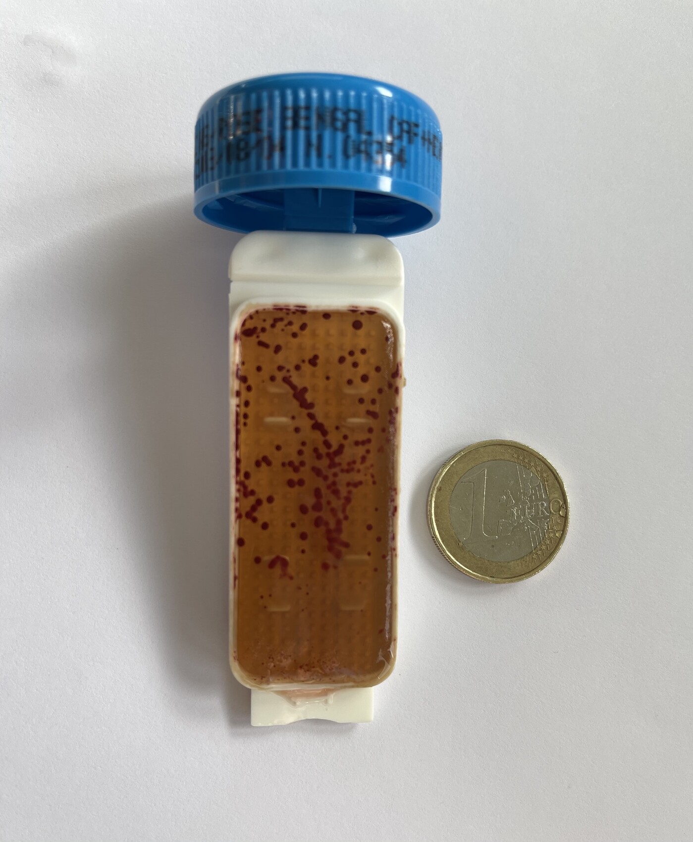 Offene Medikamentenflasche mit brauner Flüssigkeit und roten Punkten neben einer 1-Euro-Münze.