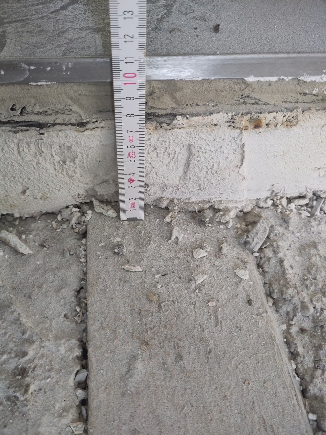 Maßband steht senkrecht an einer Wand, Messwert ca. 10 cm, daneben Beton und Geröll