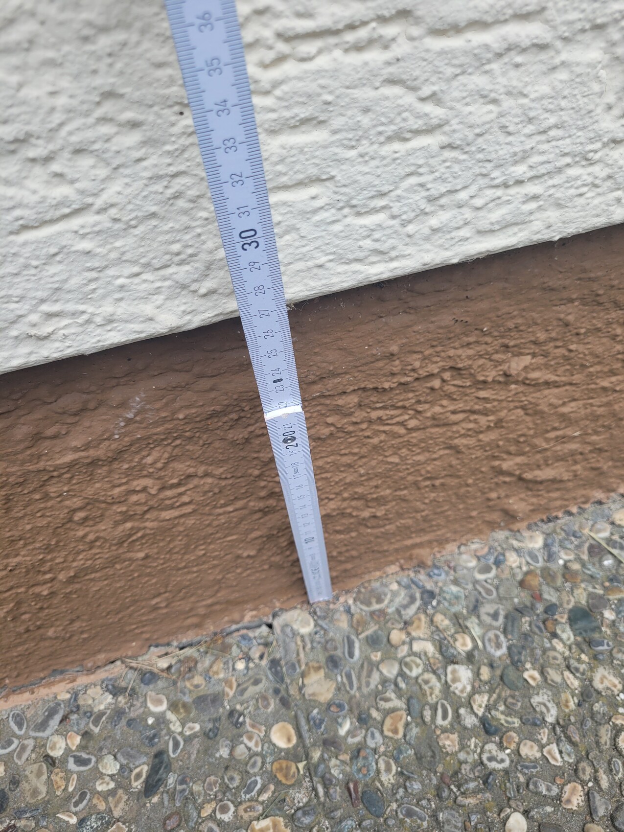 Außenwand mit weißem Putz über braunem Sockel; diagonales Maßband, Kieselboden darunter.