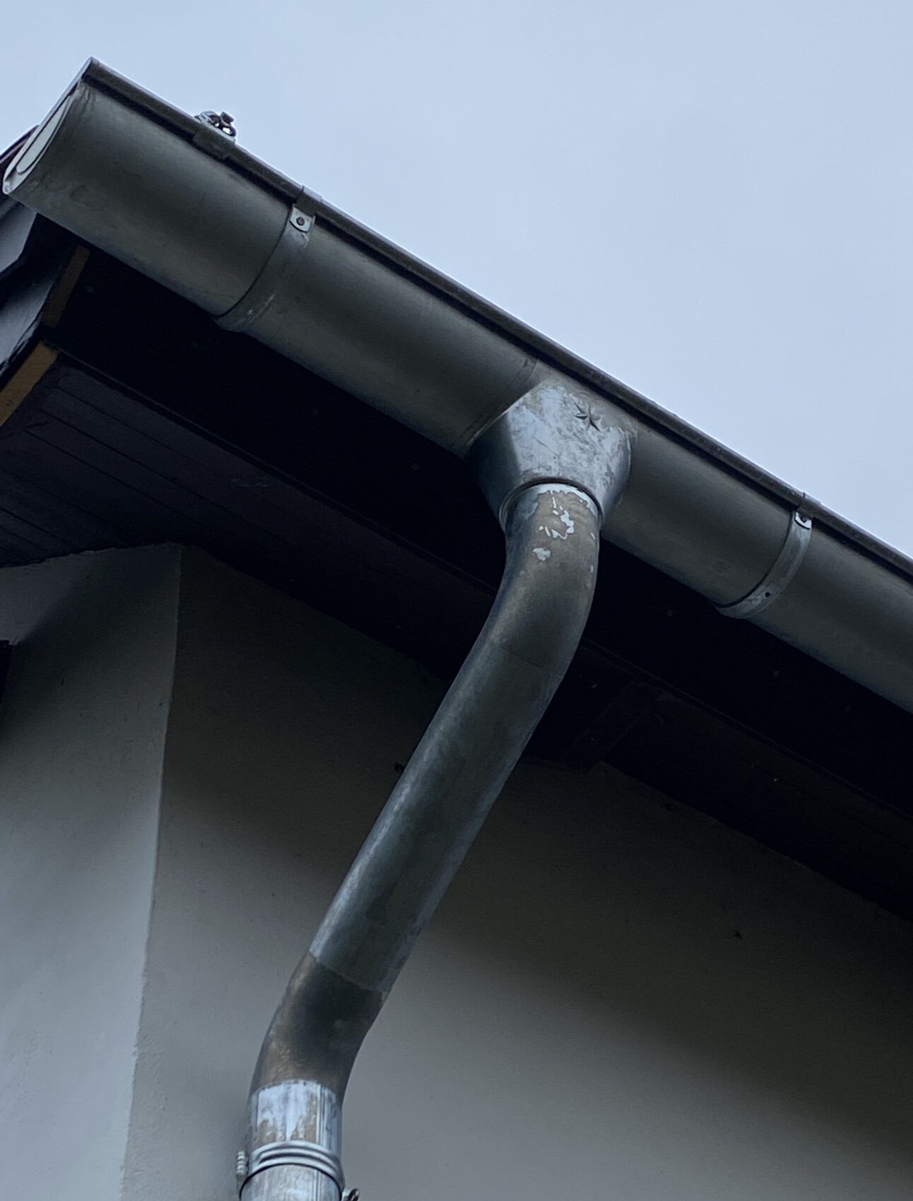 Nahaufnahme eines grauen Fallrohrs, das aus einer Regenrinne am Haus führt, blauer Himmel