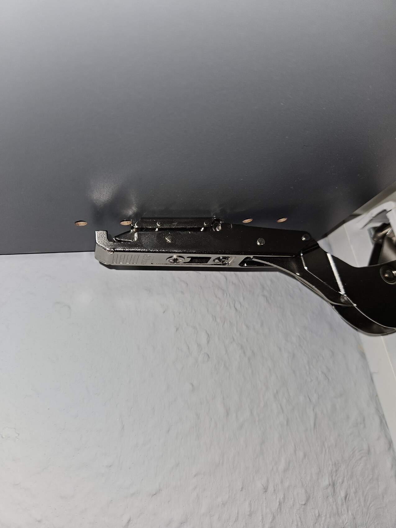 Unterseite einer grauen Platte mit schwarzem Metall-Scharnierarm und Schrauben
