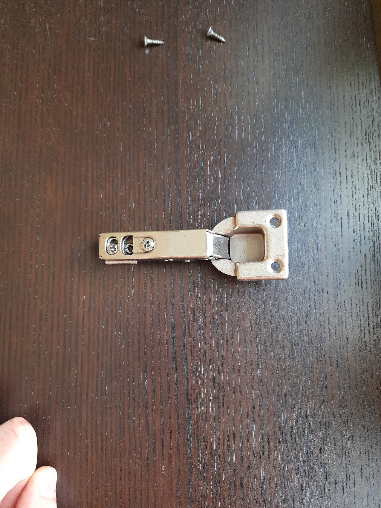 Kleines Metall-Scharnier-Bauteil liegt flach auf dunklem Holztisch; zwei Schrauben sichtbar.