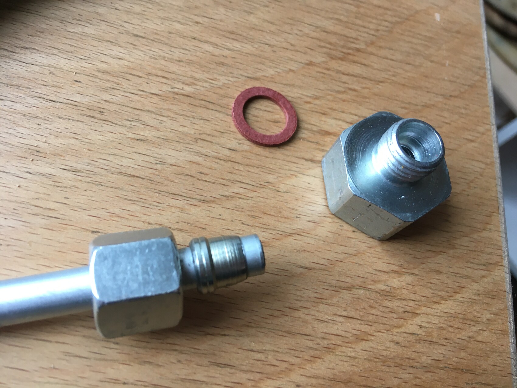 Zwei Metallfittings mit Gewinde und ein roter Dichtungsring liegen auf einem Holztisch.