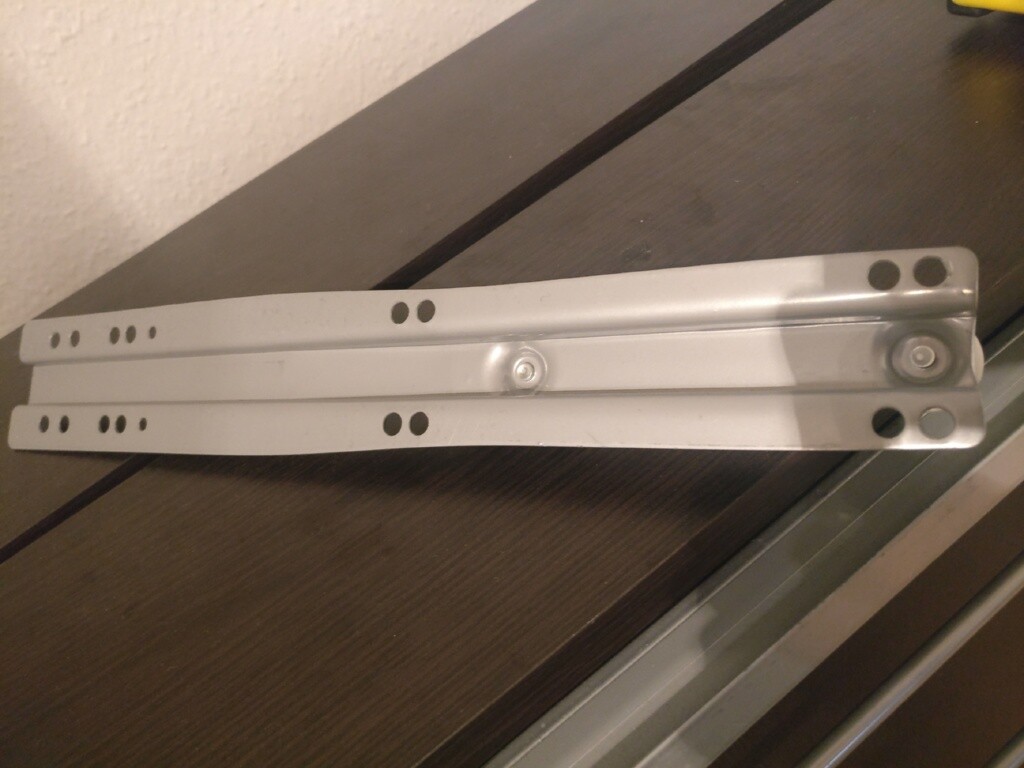 Zwei silberne Schubladenführungen mit Löchern liegen auf einer Holztischplatte.