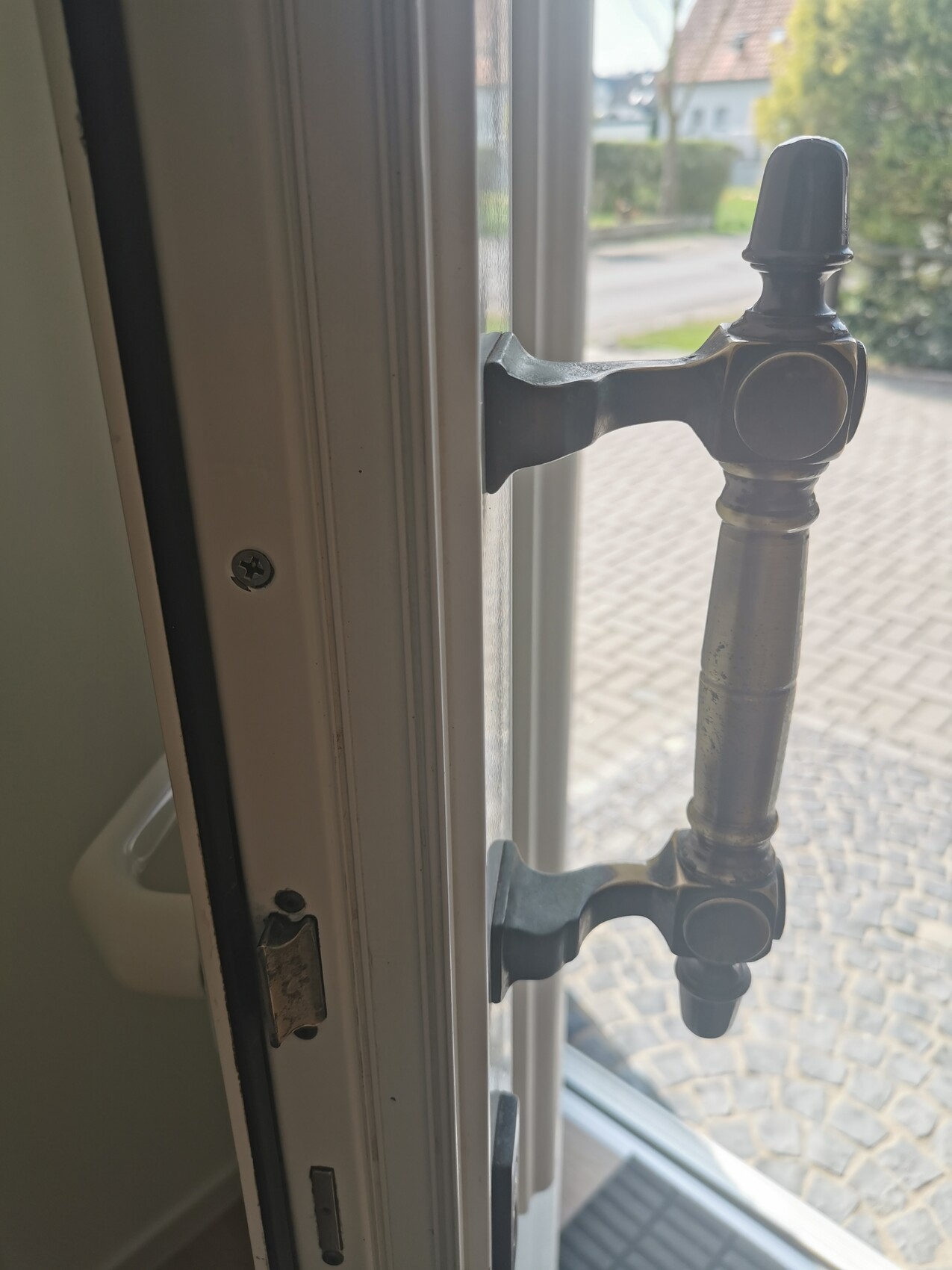 Nahaufnahme einer Espagnolette-Türverriegelung an weißem Türrahmen; draußen Straße sichtbar.