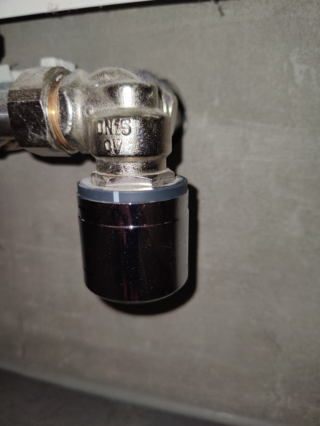 Nahaufnahme eines Metallrohrverbinders mit Ventil und schwarzem Zylinder, Betonwand im Hintergrund.