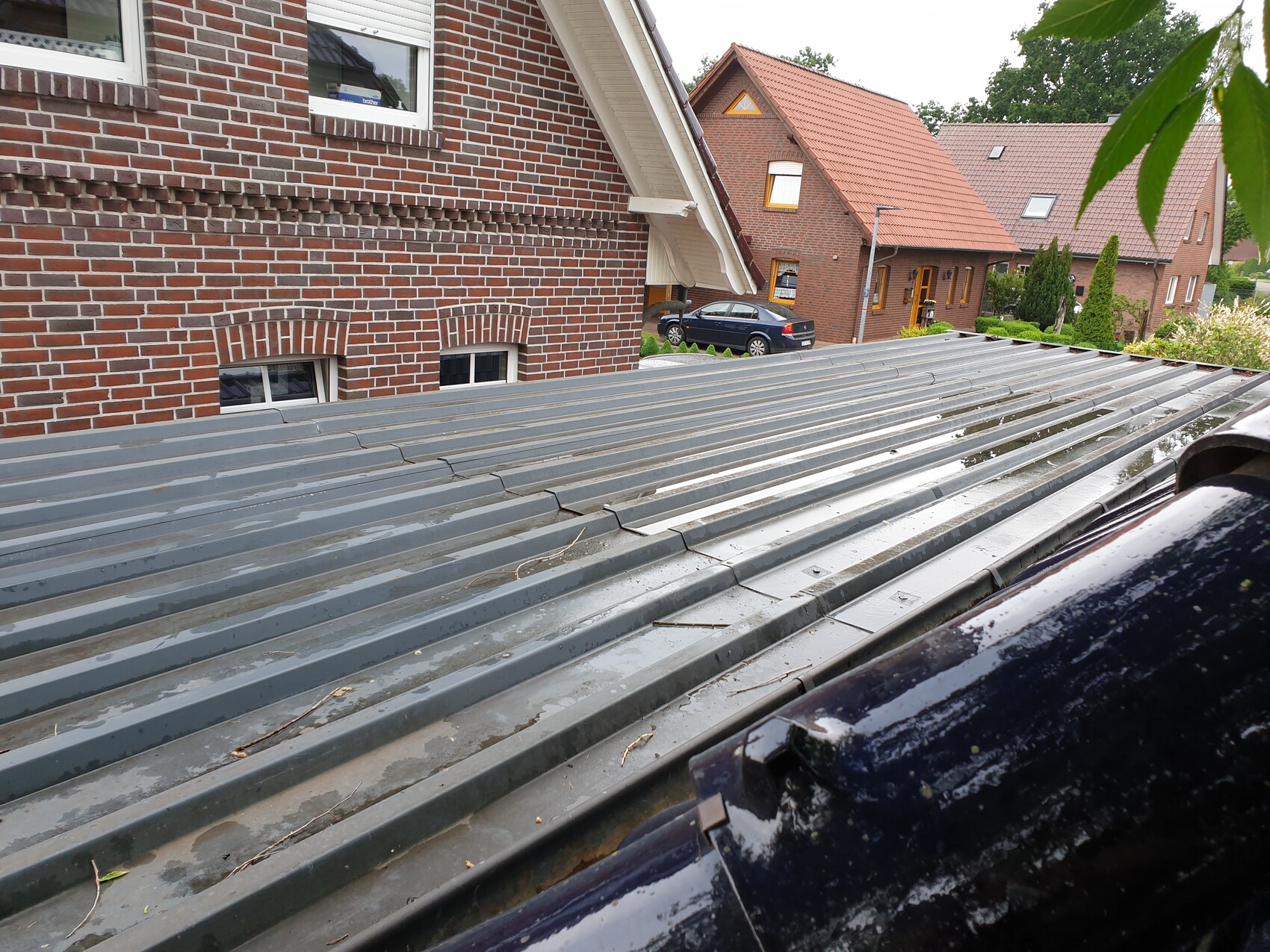 Graues Metalldach im Vordergrund, rote Backsteinhäuser und geparkter Wagen im Wohnviertel.