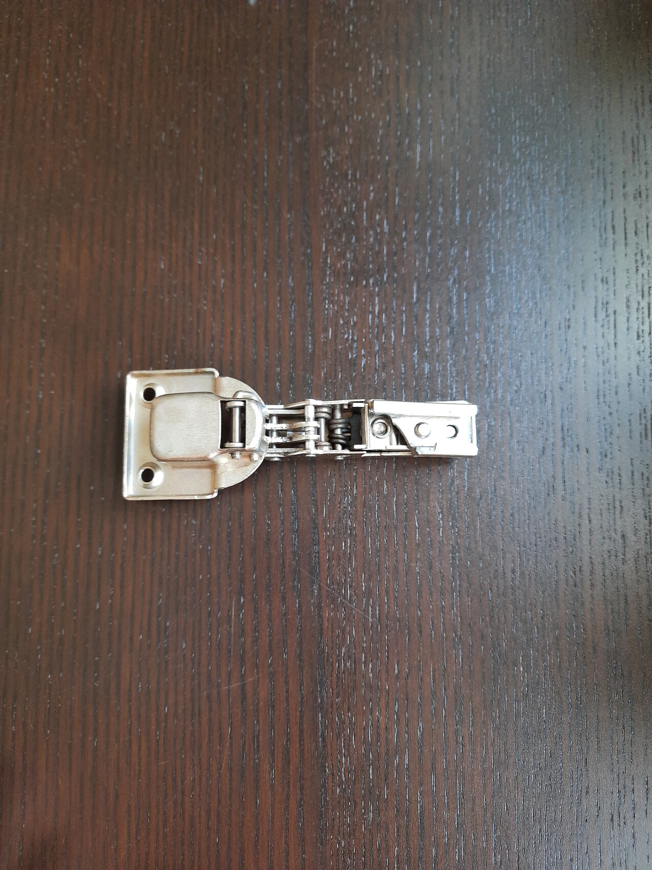 Kleines Metall-Scharnier mit Federmechanismus liegt auf dunklem Holztisch.