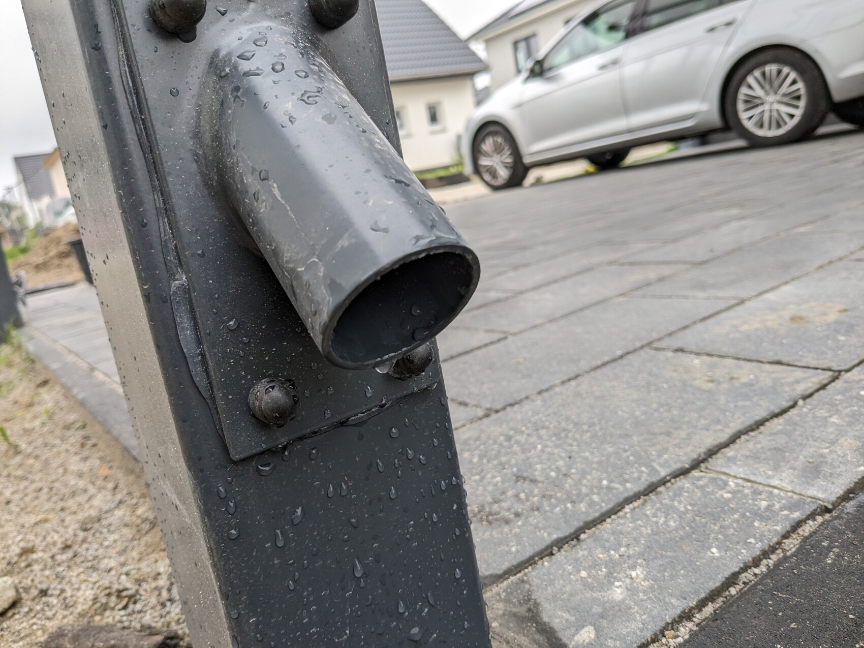 Nahaufnahme eines regennassen Metallpfostens mit gebogenem Rohr; Gehwegplatten und Auto im Hintergrund