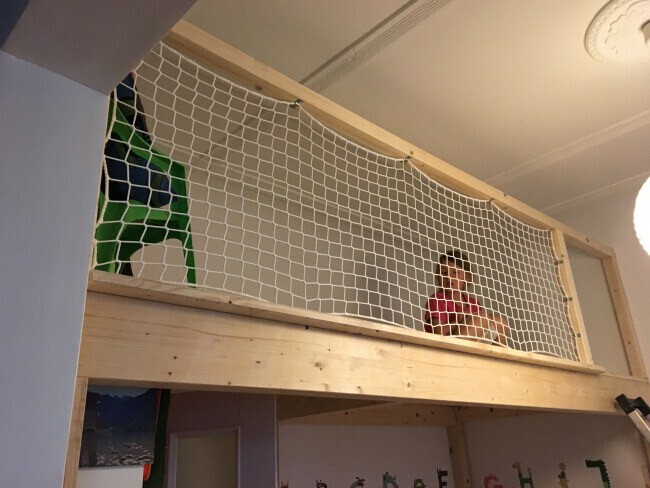 Zwei Kinder hinter einem Schutznetz an einer Holz-Loftkonstruktion im Innenraum.