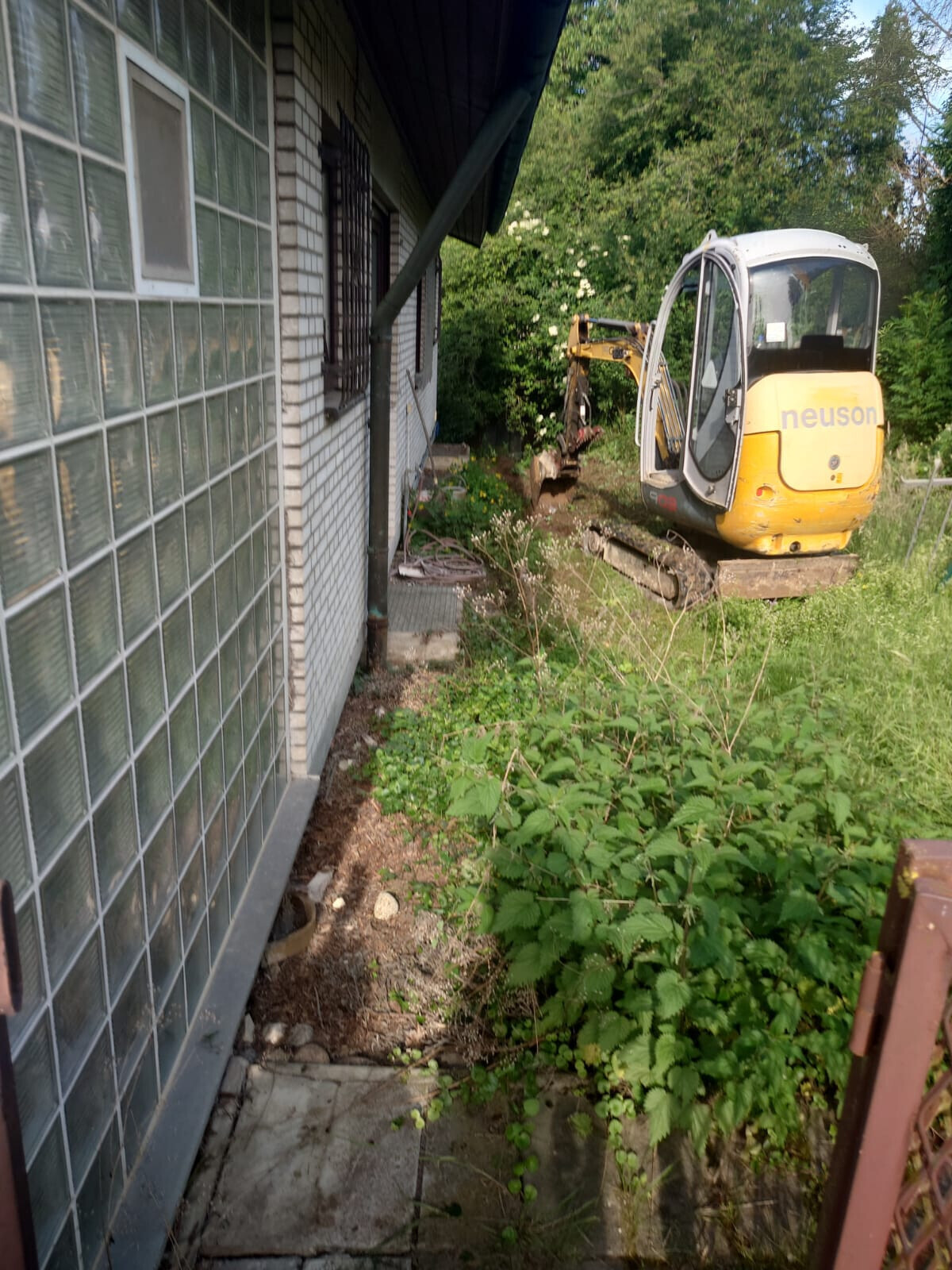 Kleine Baustelle neben Haus mit gelbem Minibagger und überwucherndem Garten