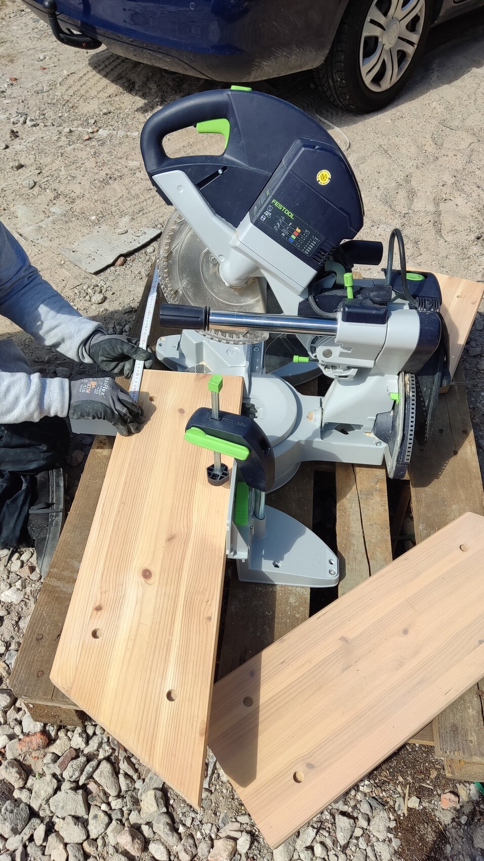 Gehrungssäge mit Holzplatten, Klemmen und grünen Griffe; Arbeiter misst Länge mit Maßband.
