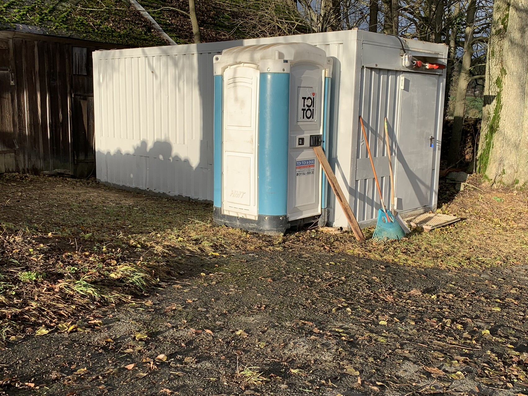 Außenansicht einer TOI TOI Porta-Potty neben einem weißen Container, mit Besen davor.