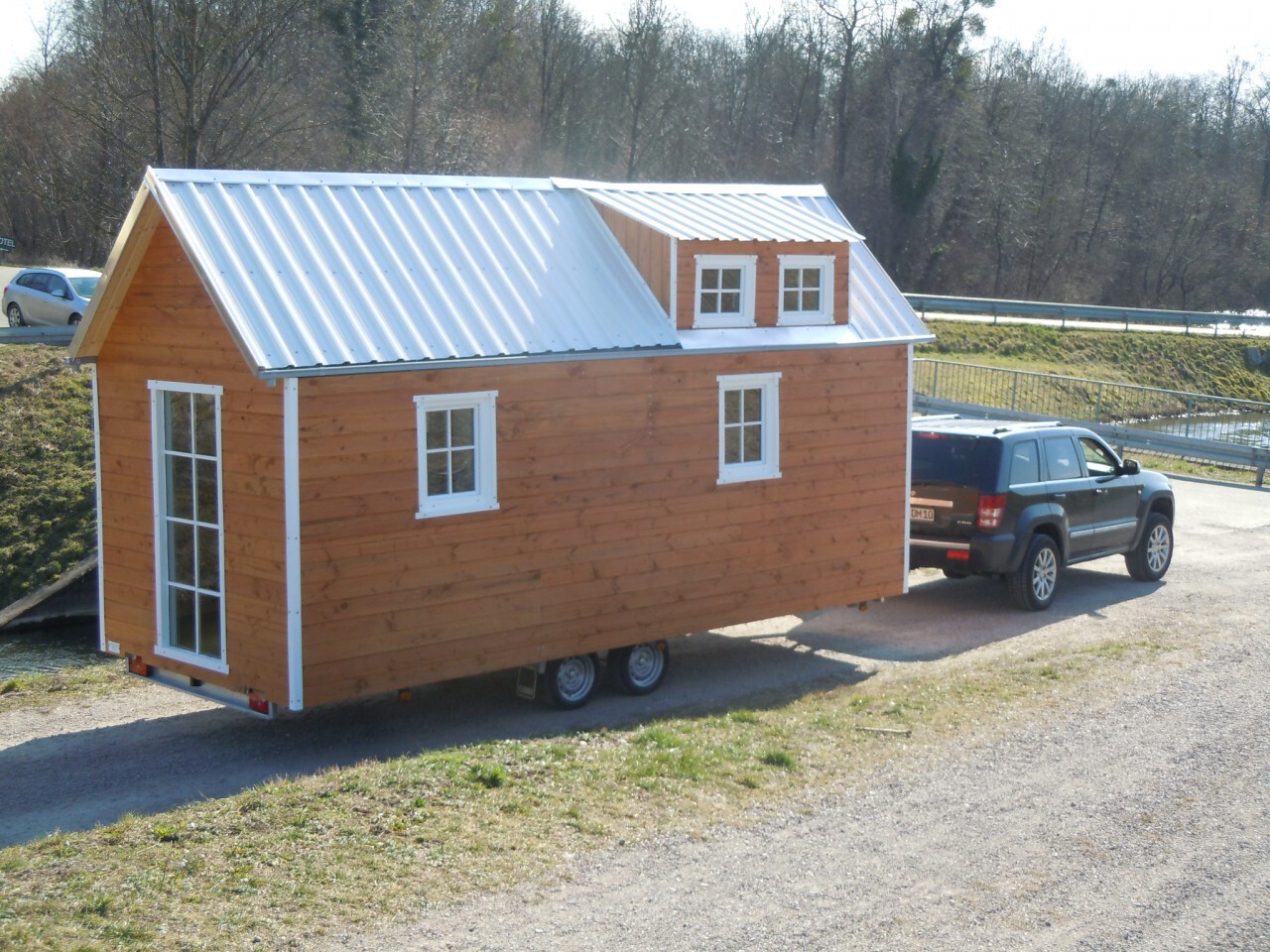 Kleines Holzhaus auf Anhänger wird von SUV gezogen; Metalldach, weiße Fenster, Wald im Hintergrund.