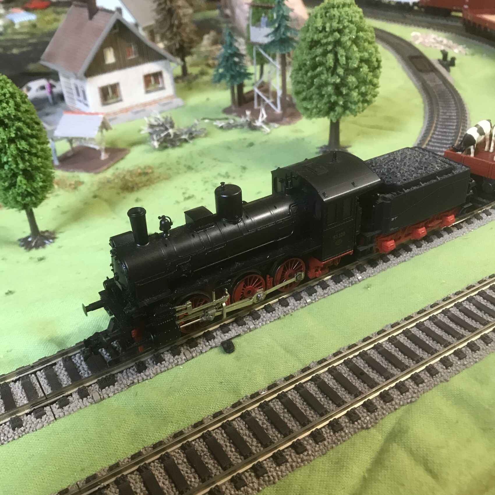 Schwarze Modell-Dampflok mit rotem Fahrwerk zieht Kohlewagen auf Modellbahn durch Landschaft.