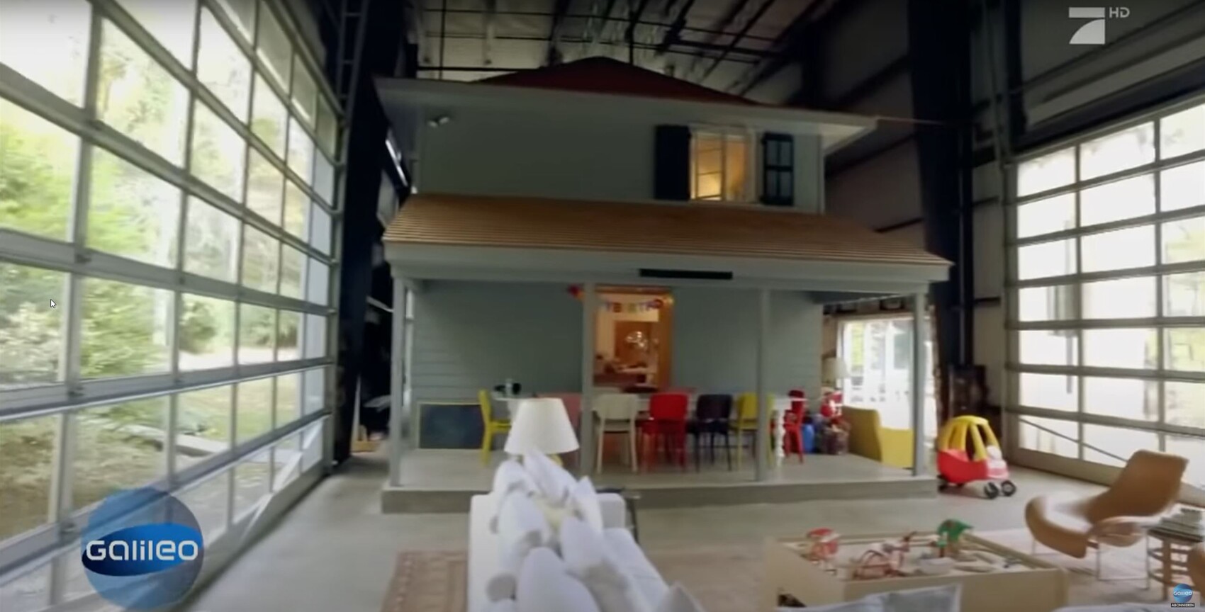 Modellhaus mit Veranda in offener Halle, umgeben von bunten Stühlen und Lounge-Möbeln.