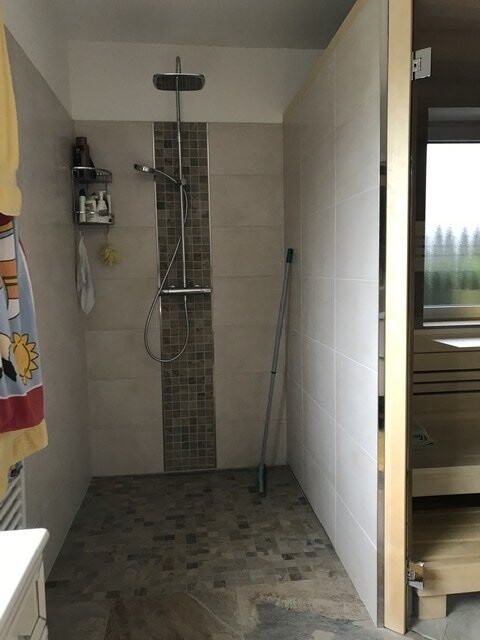 Begehbare Dusche mit Regenkopf, Handbrause, vertikaler Mosaikfliesenstreifen; Mopp lehnt an Wand.