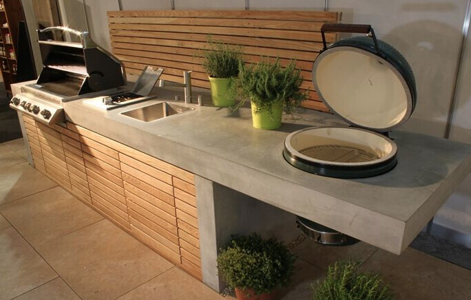 Außenküche mit Spüle, Holzfronten, Grill mit offenem Deckel und grüne Topfpflanzen daneben.